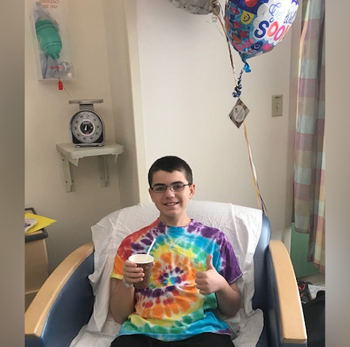一个穿着扎染t恤的男孩坐在医院的病床上，竖起大拇指。